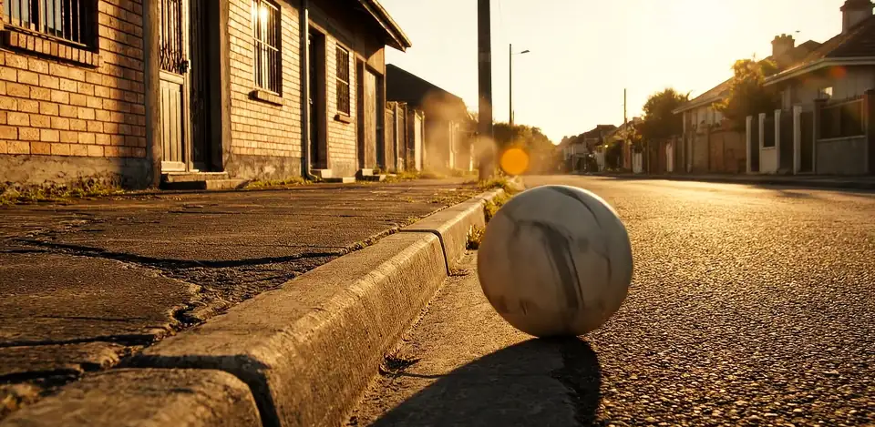 An old, worn football at rest against a kerb on an empty street at low sun.