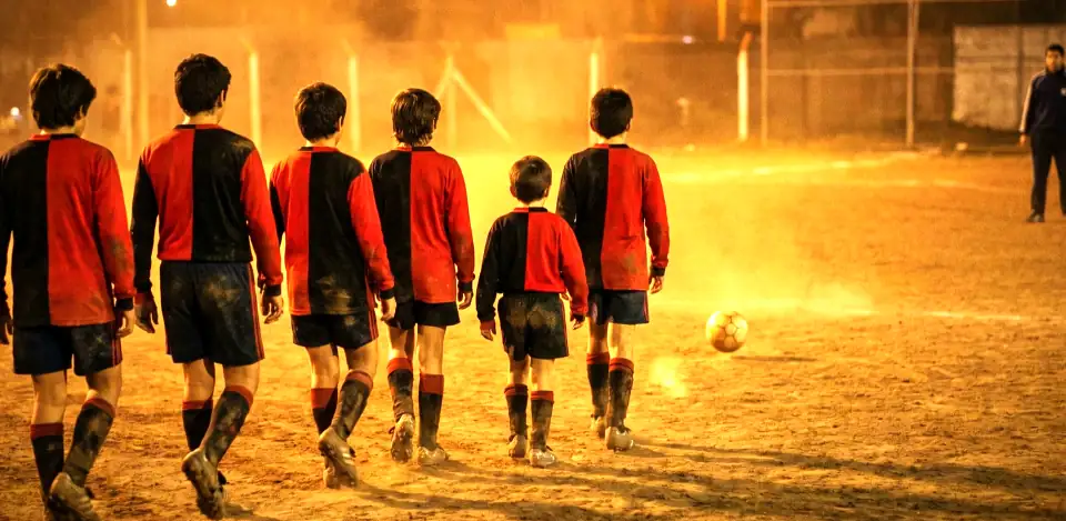 Six boys in red and black kit, seen from behind, walking across a dusty floodlit pitch behind a ball.