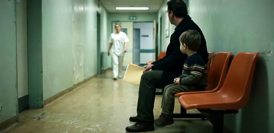 A man and a small boy, seen from behind, waiting on orange chairs in a pale green clinic corridor.