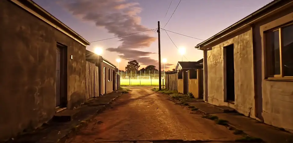 A dirt lane between low houses at dusk, leading to a lit pitch.