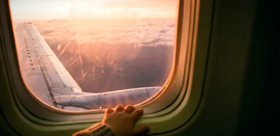A hand on an aeroplane window, with the wing and pink clouds outside.