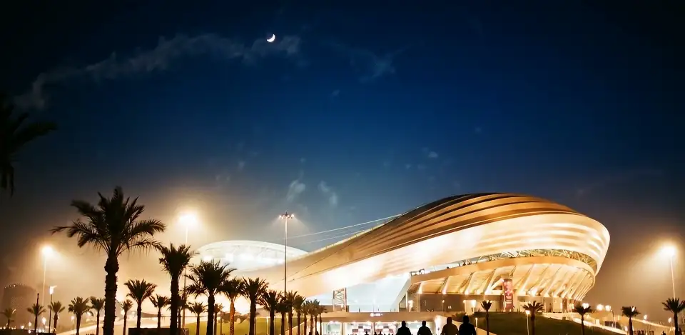 Figures silhouetted walking towards a glowing gold-roofed stadium among palm trees, under a dark blue sky with a crescent moon.