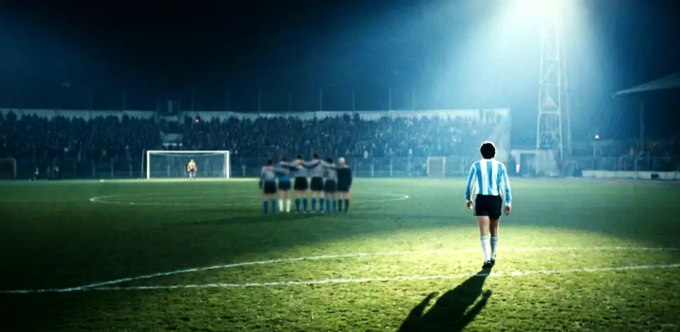A lone player in pale blue and white stripes walking towards a distant goal and a huddled team, on a blue-lit pitch at night.