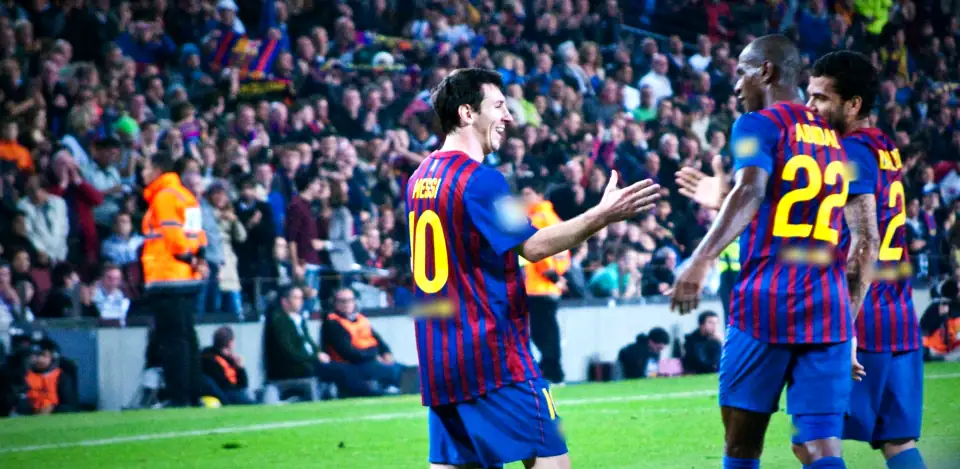 Messi, smiling, reaching out to two Barcelona teammates on the pitch, with a full stand behind them. Crests and sponsors blurred in the film.