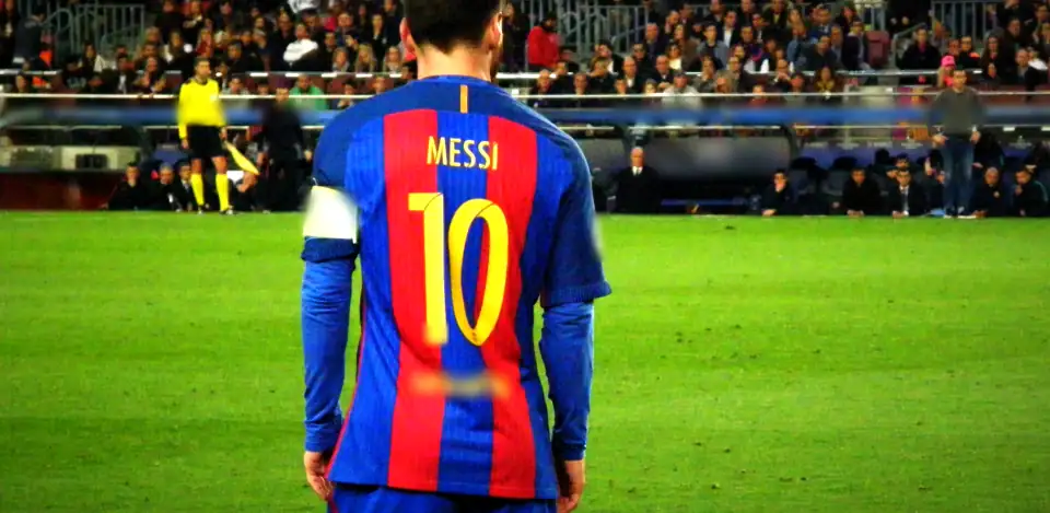Messi seen from behind in a Barcelona shirt with a yellow 10, stands and a linesman ahead. Sponsor blurred in the film.