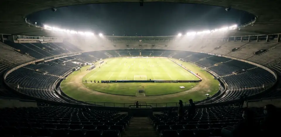 A huge dark oval stadium with empty tiers and a bright floodlit pitch at the centre.