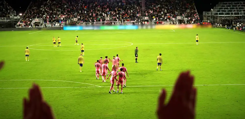 Inter Miami players in pink gathered on the pitch at night, visiting players in yellow around them, raised hands silhouetted in the foreground. Inter Miami v Real Salt Lake. Boards blurred in the film.
