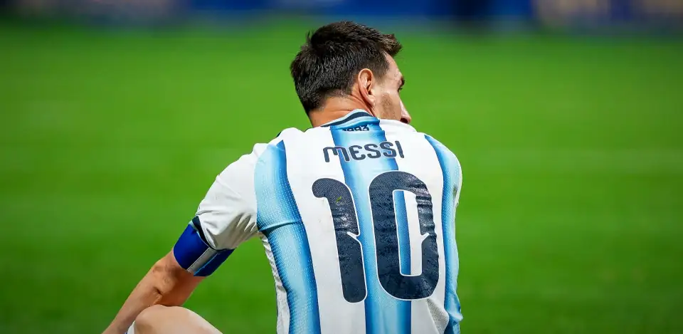 Messi seated on the grass, seen from behind and turning his head, in Argentina's 10 shirt, against plain green. Argentina v Egypt, 7 July 2026.