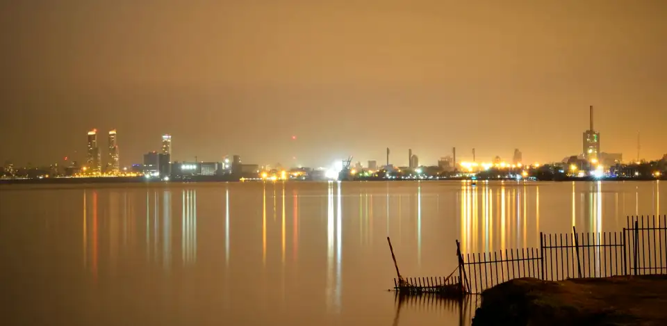 The Rosario riverfront at night, with lights across the water under an orange sky and a broken fence in the foreground.