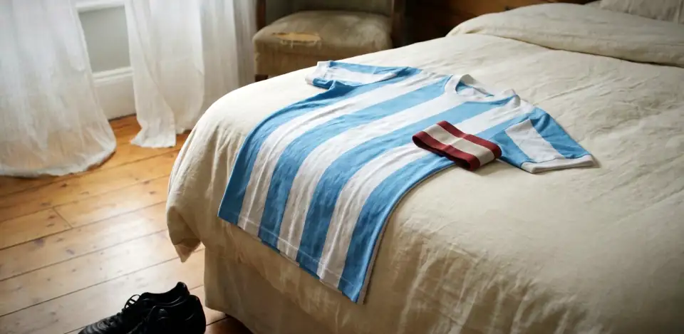 A striped pale blue and white shirt and a folded armband on a bed in a sunlit room, with boots on the floorboards.