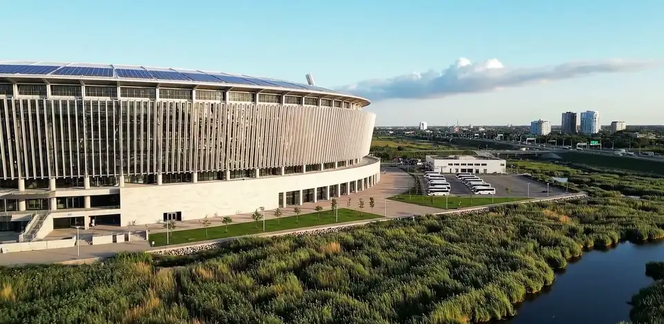 Slow aerial move past a modern stadium, wetlands and blue sky.