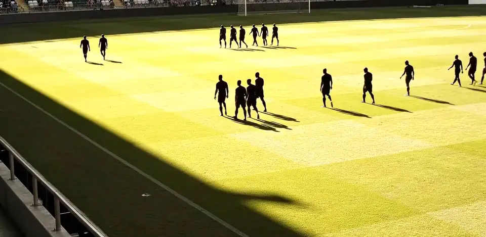 Small black figures walking across a sunlit pitch, a long stand shadow in the foreground.