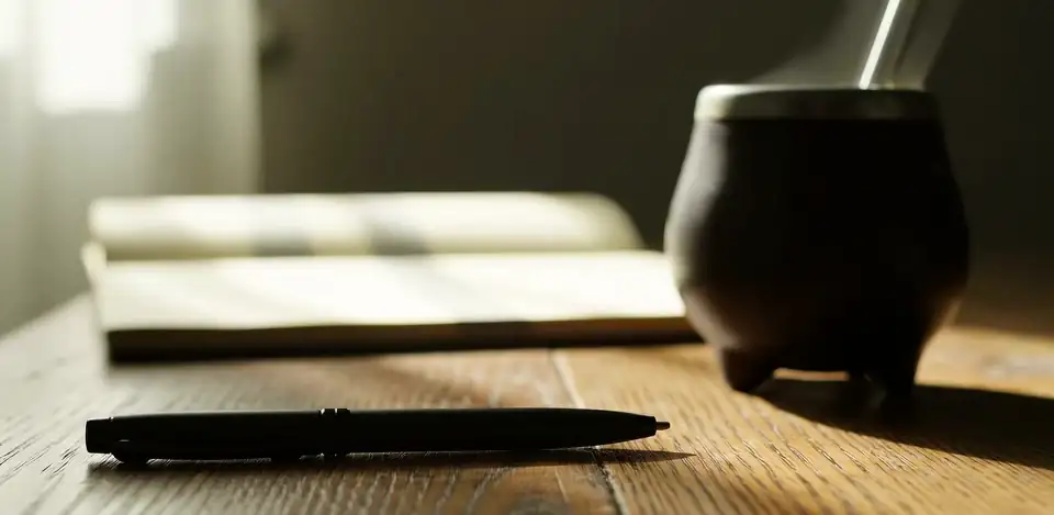 Steam rising from a mate gourd, with an open book and a pen on a wooden desk in soft light.